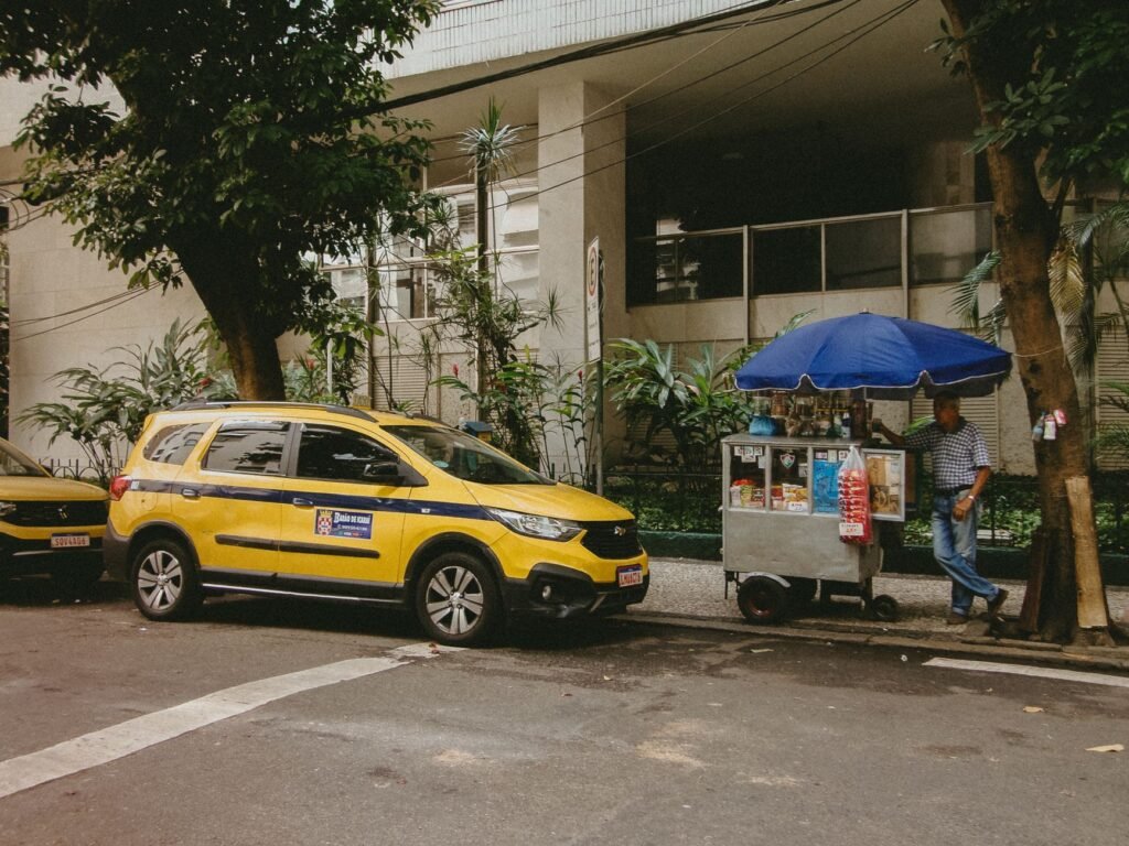 Uma cena típica de rua no Rio de Janeiro, mostrando um táxi amarelo e um vendedor ambulante em frente a um prédio moderno.