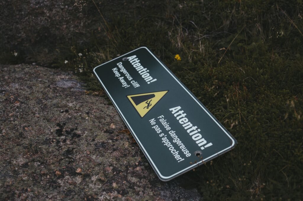 Uma placa de aviso ao ar livre dizendo 'Atenção! Penhasco perigoso. Mantenha distância.' caída em um chão rochoso.